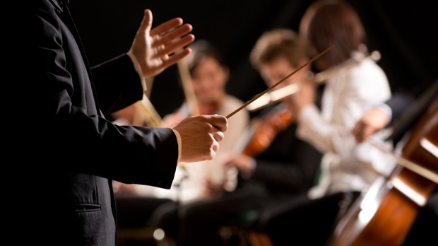 Maestro dirigindo orquestra sinfônica com artistas no fundo, mãos close-up.