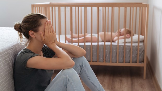 młoda matka siedzi przy łóżeczku swojego dziecka skrywając twarz w dłoniach i przeżywa depresję poporodową